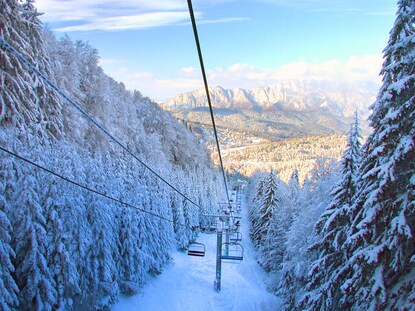  Snow covered ski lift with evergreen trees on either side and picturesque mountains in the distance ahead. 