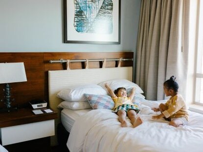 Two children smile while sitting on a comfortable bed in a Suite at The Grand Islander, a Hilton Grand Vacations Club in Oahu, Hawaii